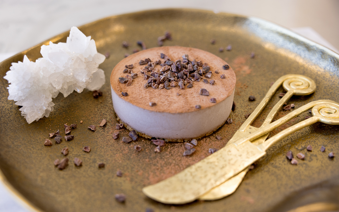 A dessert plate featuring a round chocolate mousse dusted with cocoa and sprinkled with cacao nibs, accompanied by a gold dessert fork and a cluster of white sugar crystals.