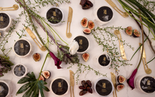 Load image into Gallery viewer, An aerial view of various wheels of SriMu vegan cheese, spiral knives, and figs, arranged amidst greenery and flowers on a marble surface.

