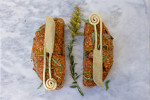 Load image into Gallery viewer, Two pieces of bread with red cheese, garnish and brass knives on top of it with flower as decor

